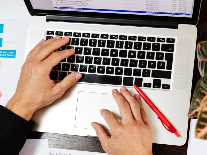 Overhead view of hands on a laptop keyboard with a red pen resting beside the trackpad